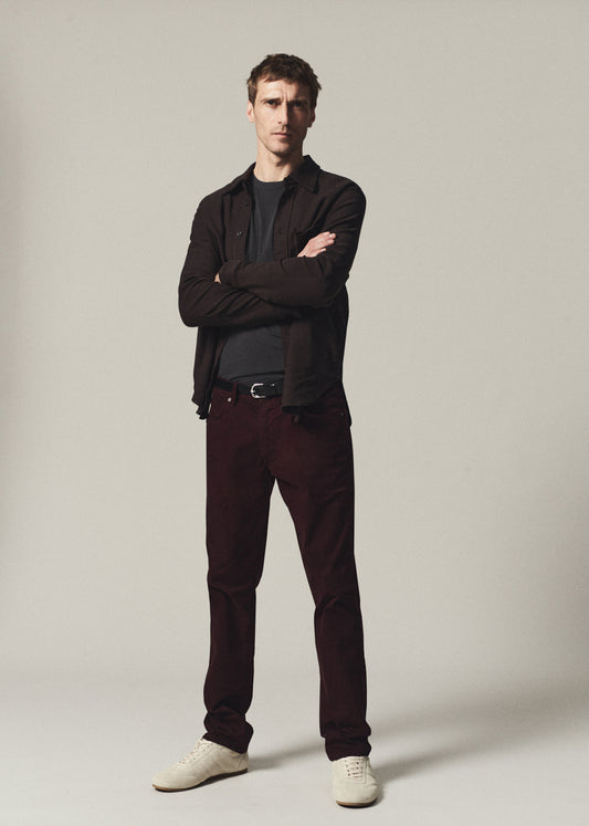 A man modeling a dark brown zip-up jacket over a black t-shirt, paired with dark burgundy corduroy trousers and white sneakers, standing against a neutral light gray background.