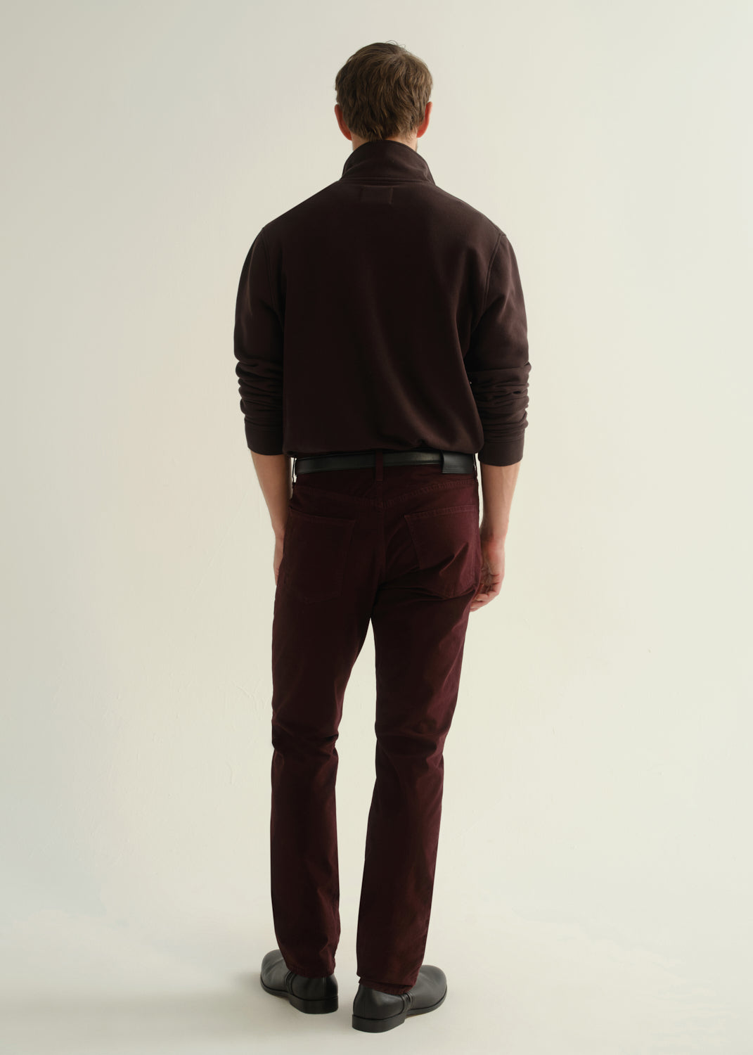 Person wearing a dark brown collared pullover sweatshirt, burgundy straight leg pants, and black leather boots, back full body view on a light grey background.