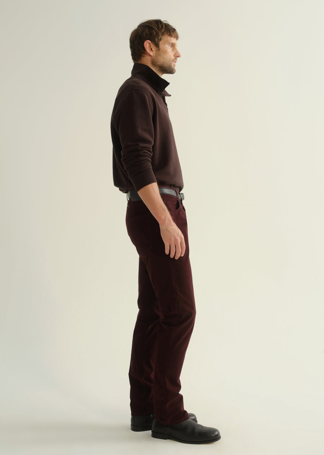 Person wearing a dark brown collared pullover sweatshirt, burgundy straight leg pants, and black leather boots, side full body view on a light grey background.
