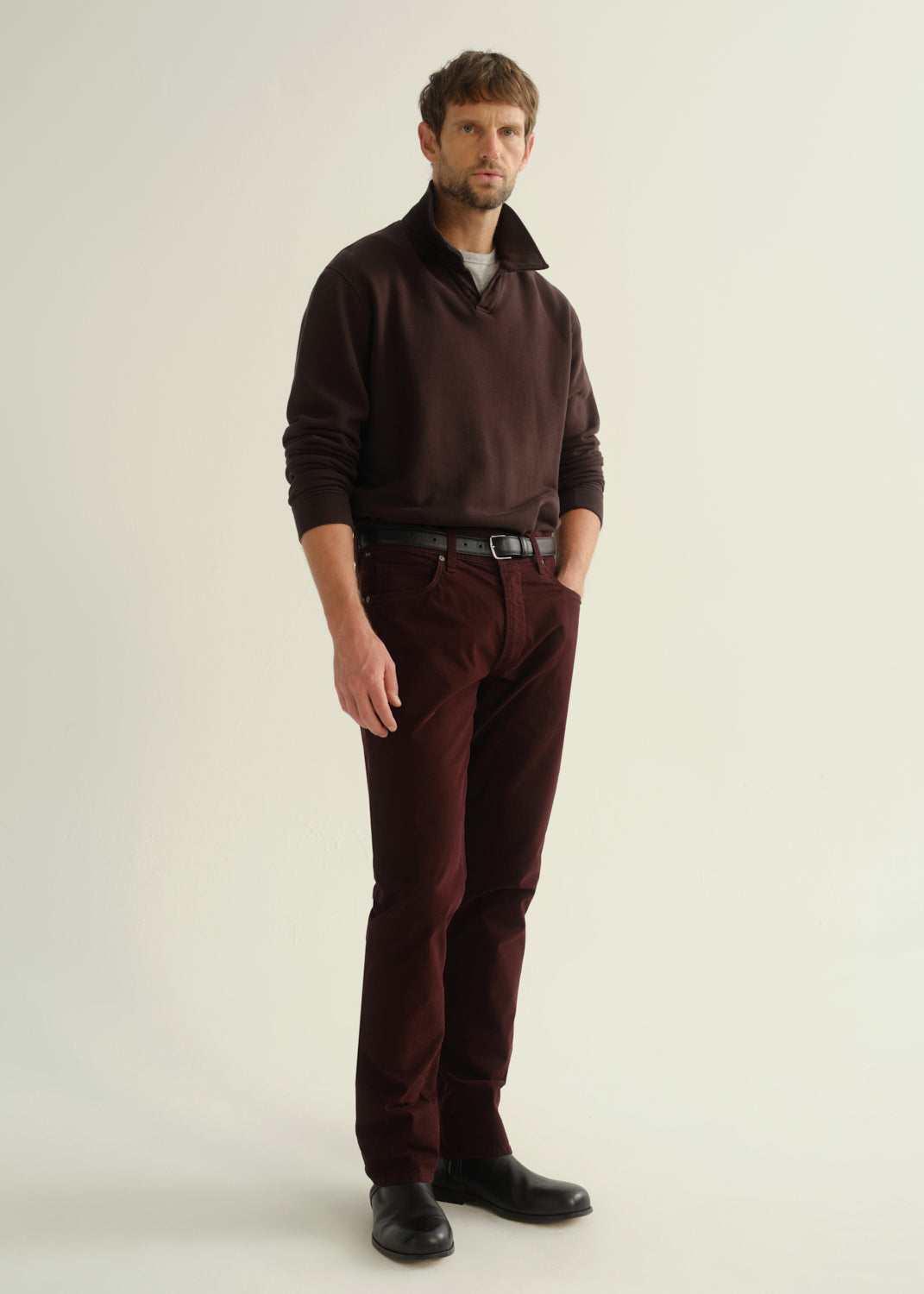Person wearing a dark brown collared pullover sweatshirt, burgundy straight leg pants, and black leather boots, front full body view on a light grey background.
