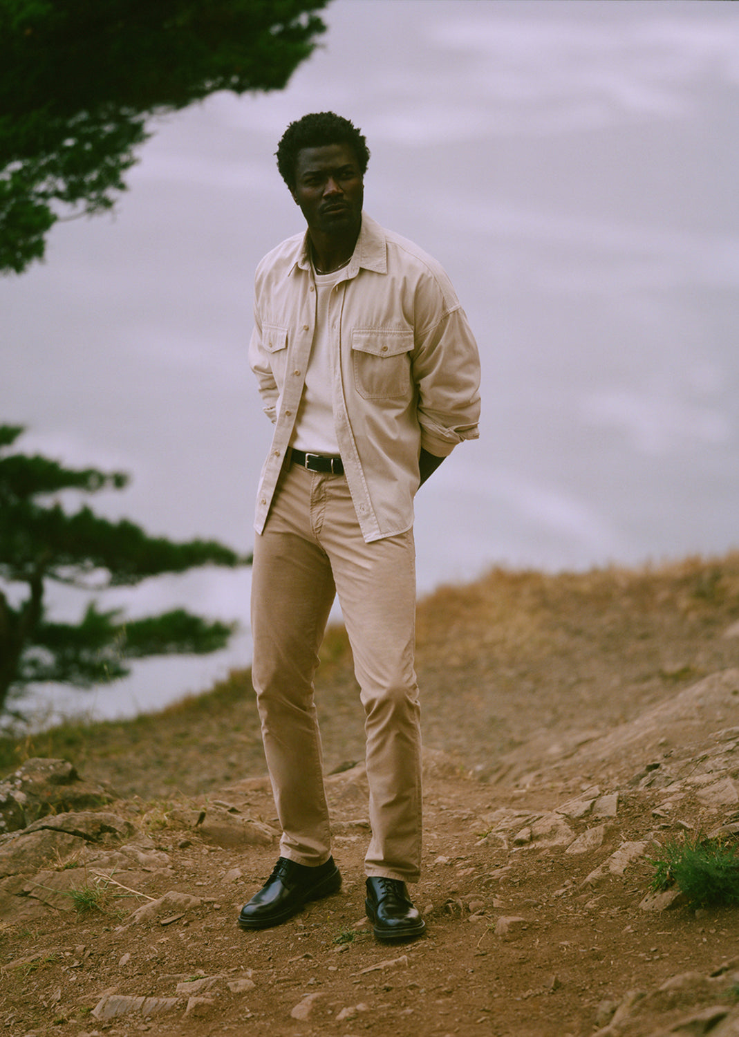 A man stands outdoors on a rocky hillside wearing a light beige overshirt, matching beige trousers, a white shirt, and black shoes, with a soft-focus natural background.