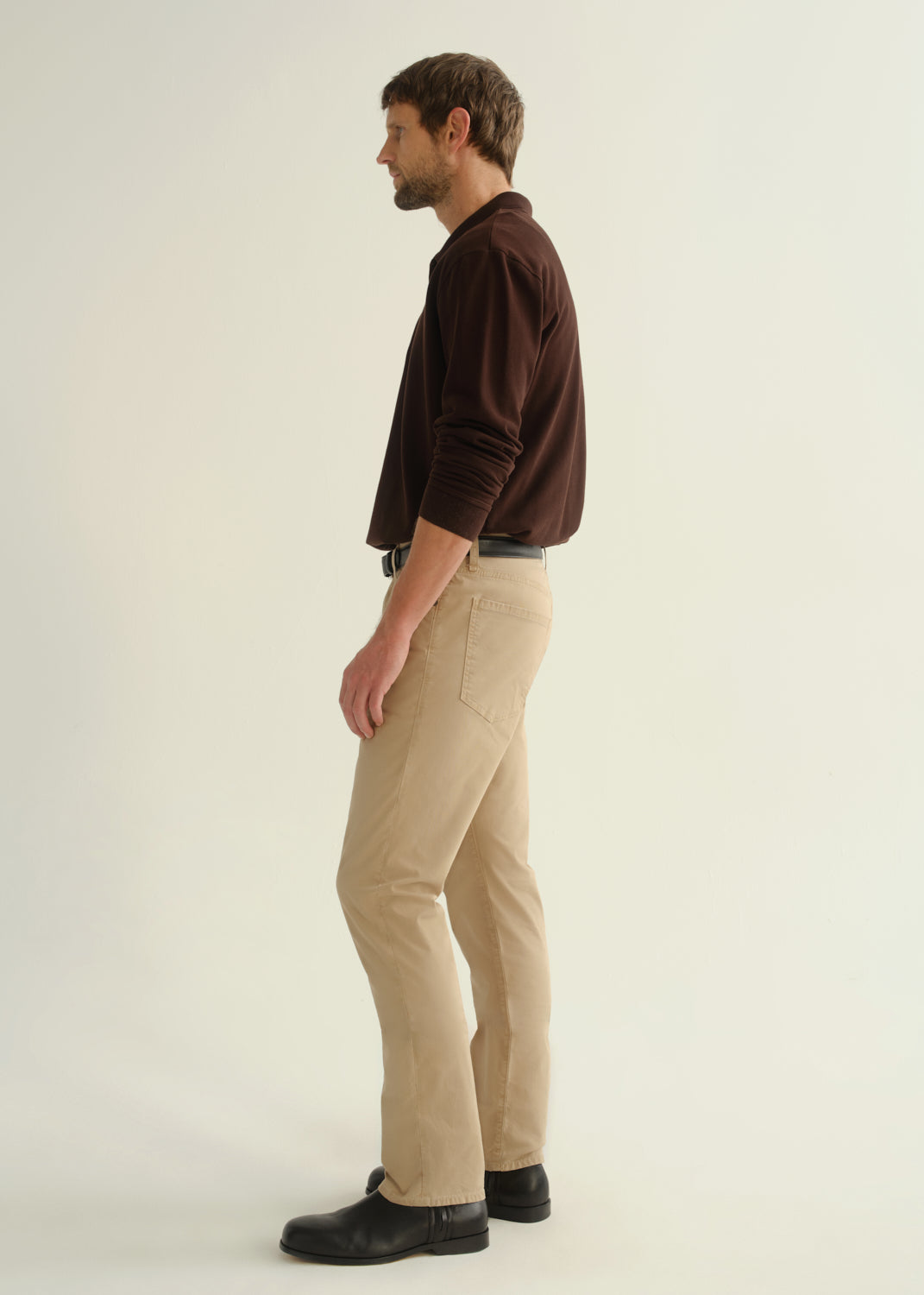 Person wearing a dark brown collared pullover sweatshirt, beige pants, and black leather boots, side full body view on a light grey background.
