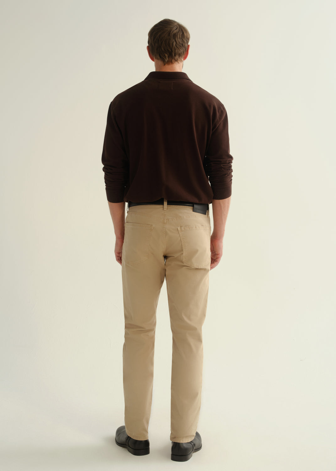 Person wearing a dark brown collared pullover sweatshirt, beige pants, and black leather boots, back full body view on a light grey background.