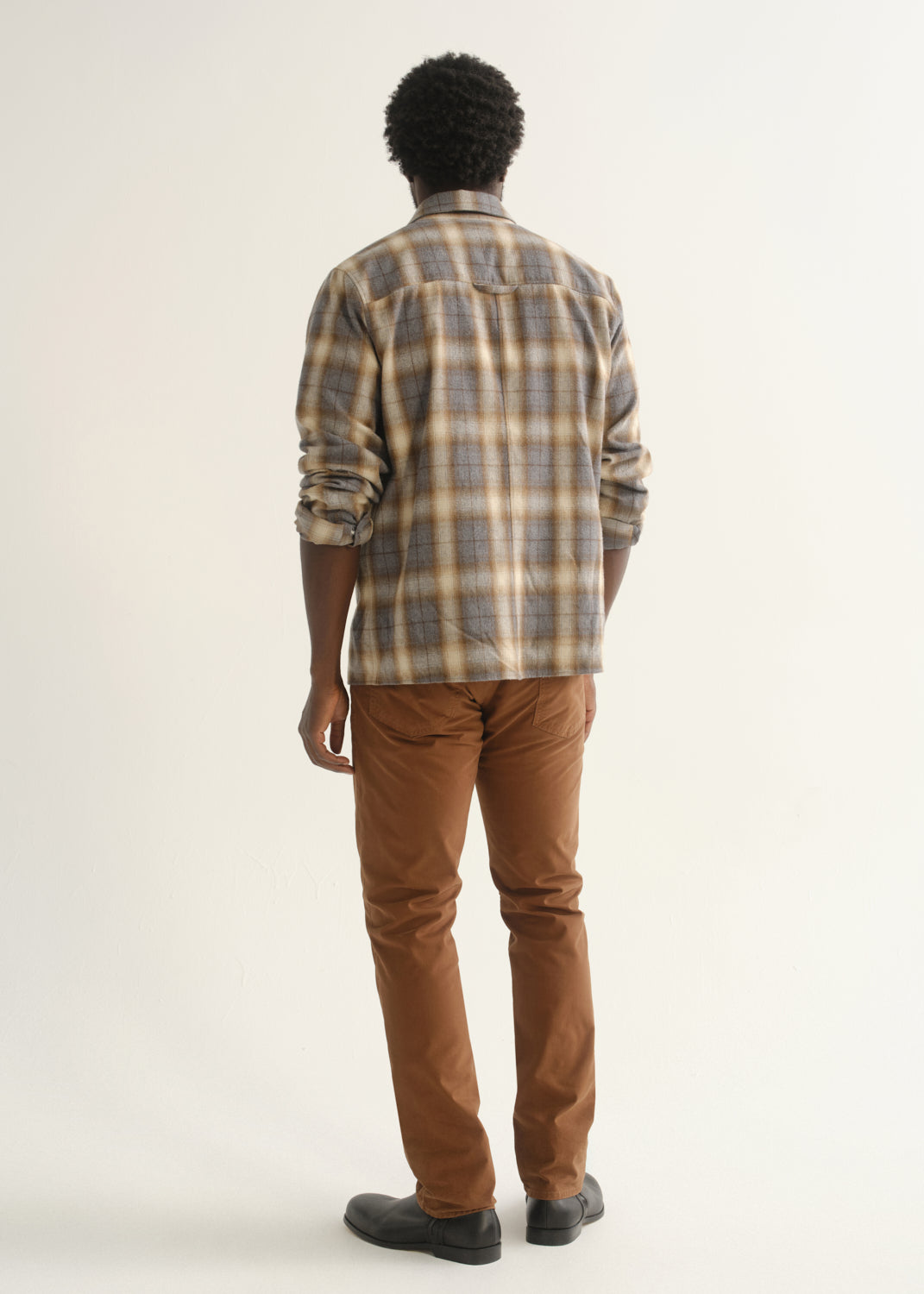 Person wearing rust brown straight leg pants with a multicolor plaid button-up shirt and black shoes, back full body view on a light grey background.