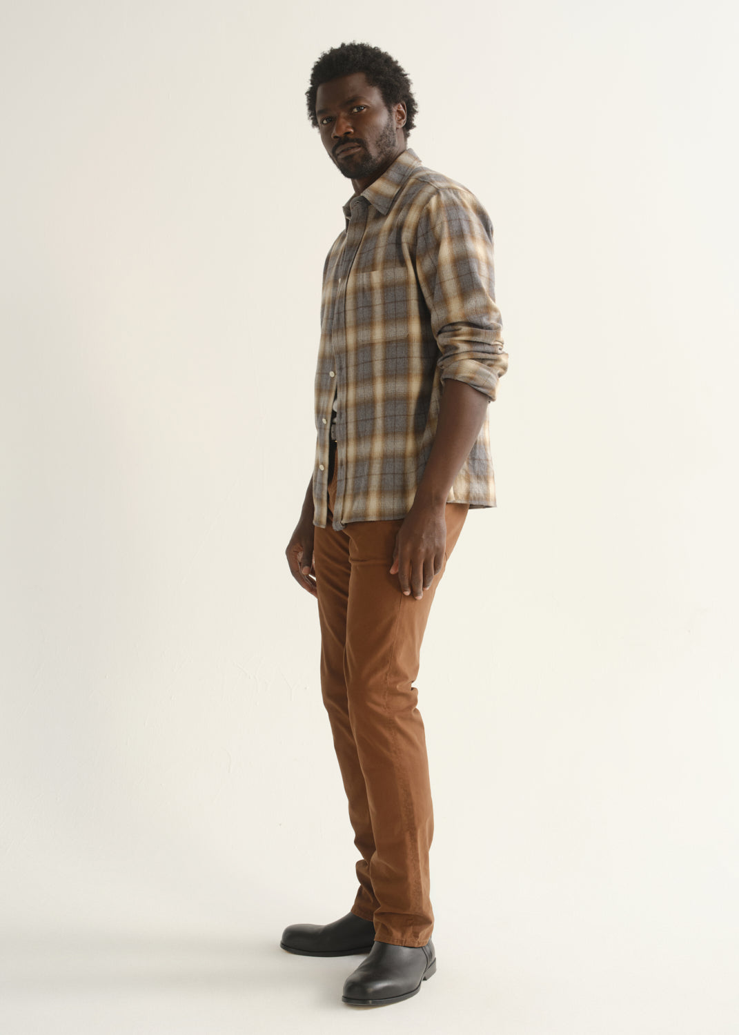 Person wearing rust brown straight leg pants with a multicolor plaid button-up shirt and black shoes, angled front full body view on a light grey background.