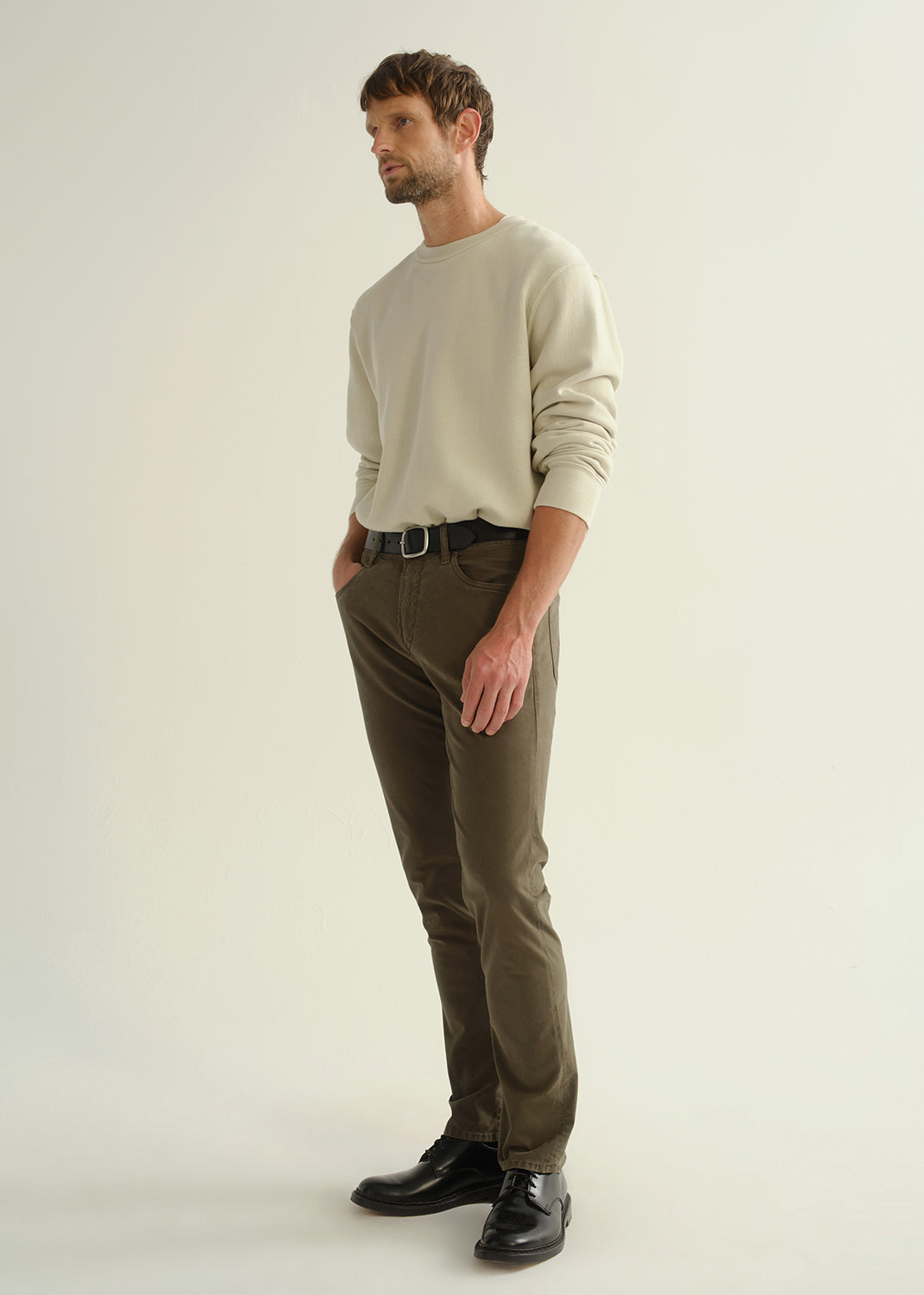 Person wearing a light grey crewneck sweatshirt, dark olive green pants, and black leather shoes, angled front full body view on a light grey background.
