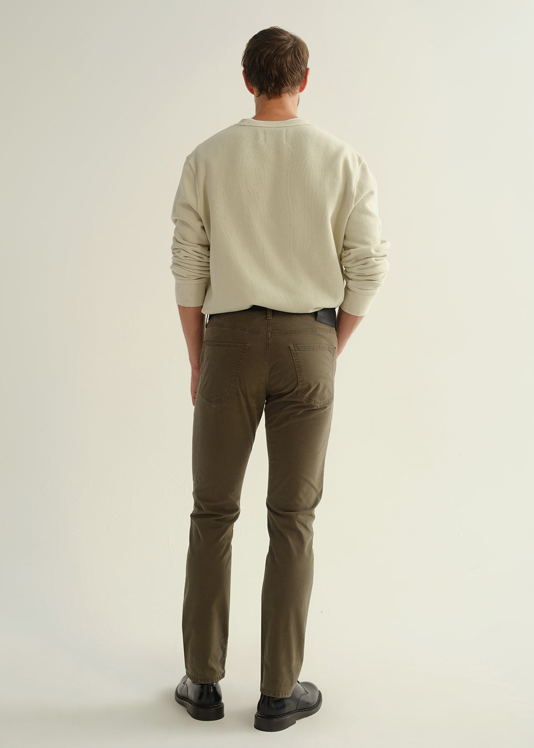 Person wearing a light grey crewneck sweatshirt, dark olive green pants, and black leather shoes, back full body view on a light grey background.