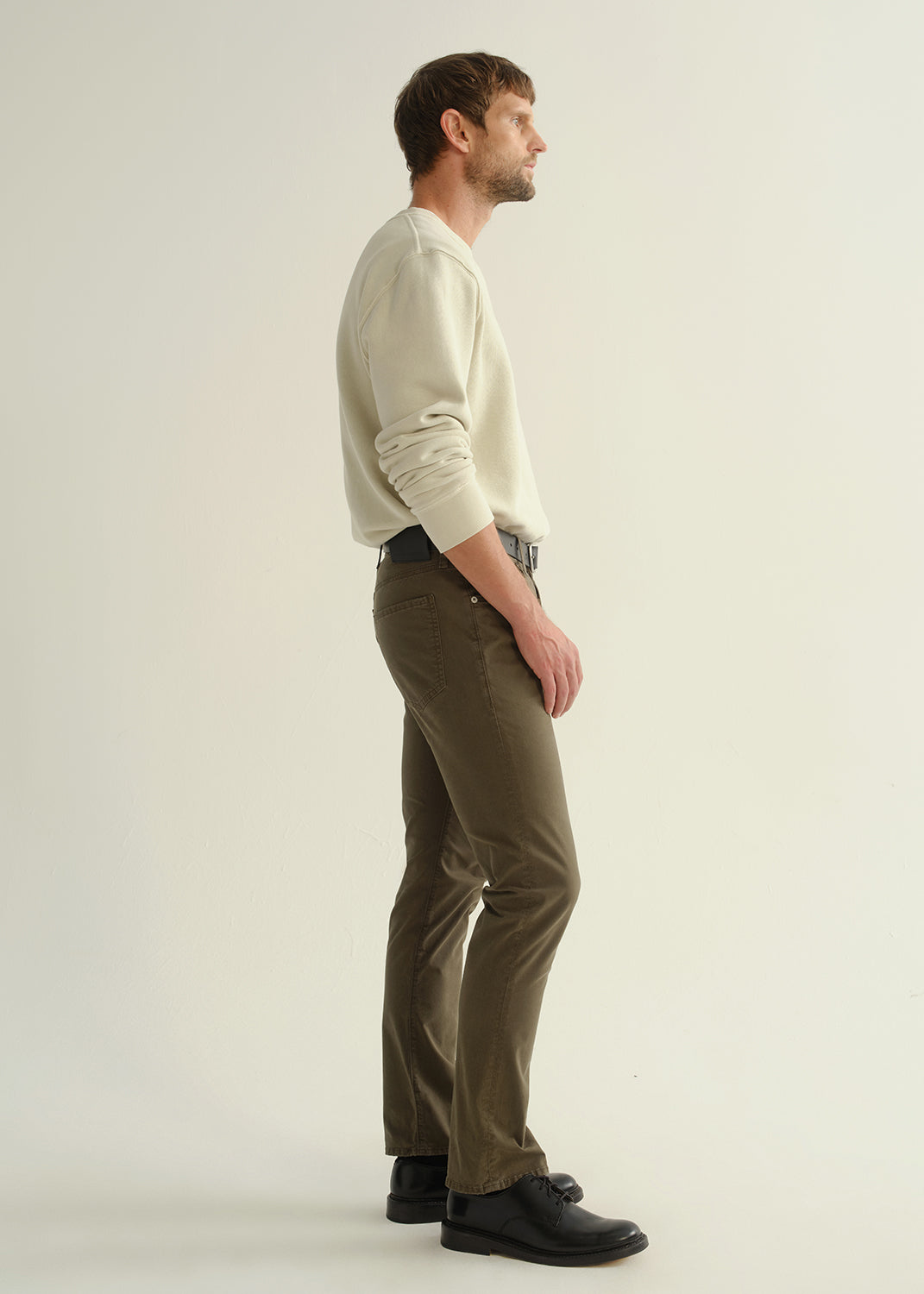 Person wearing a light grey crewneck sweatshirt, dark olive green pants, and black leather shoes, side full body view on a light grey background.