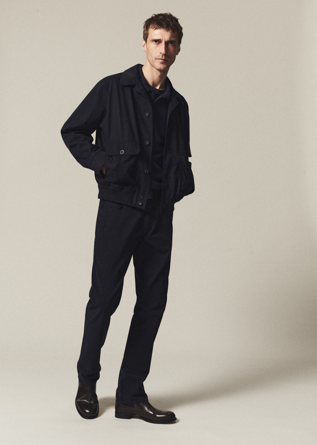 A man modeling a dark navy blue jacket and matching trousers against a plain beige studio background.