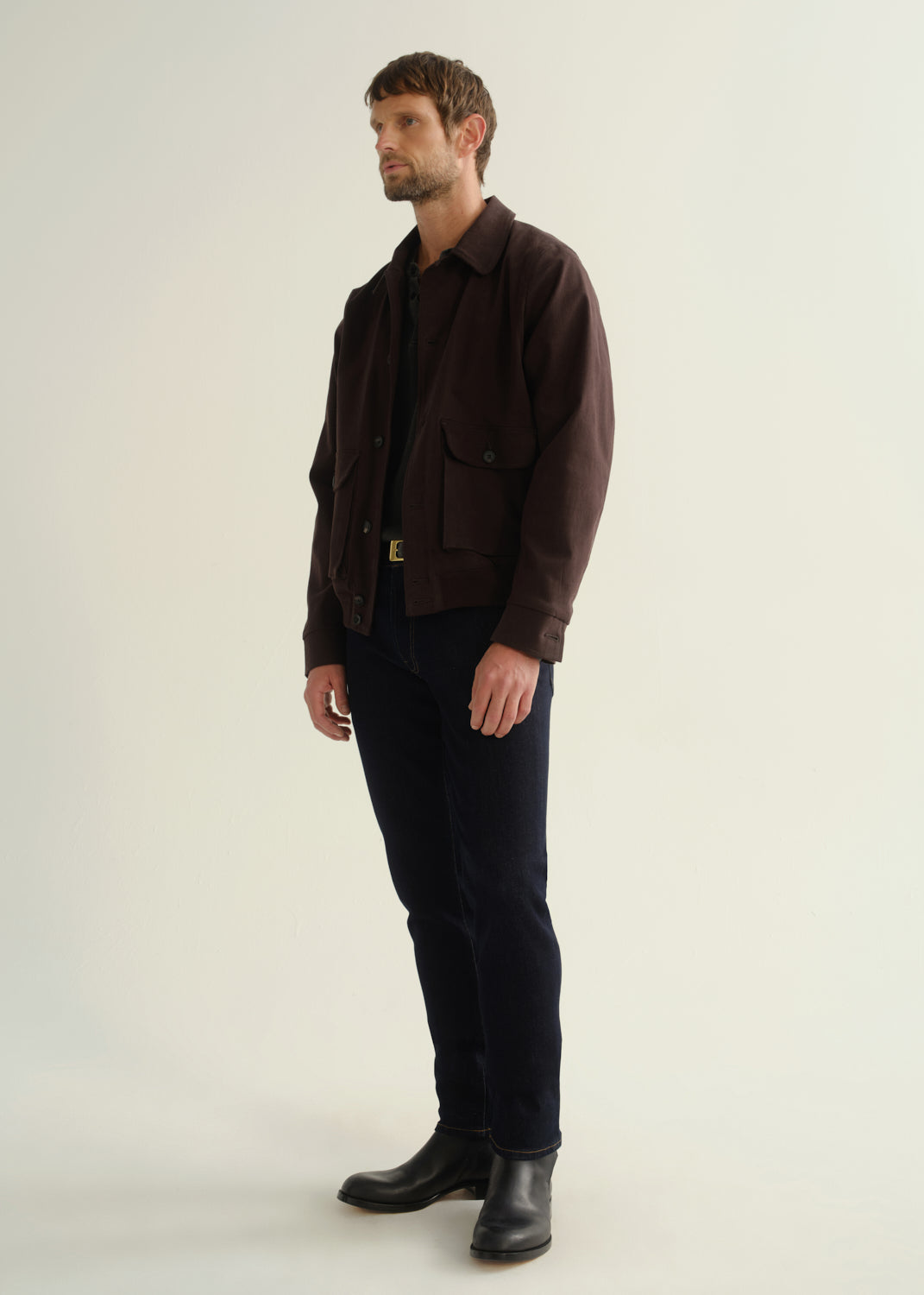 Person wearing a dark brown button-front jacket with flap pockets, dark indigo jeans, and black leather boots, front full body view on a light grey background.
