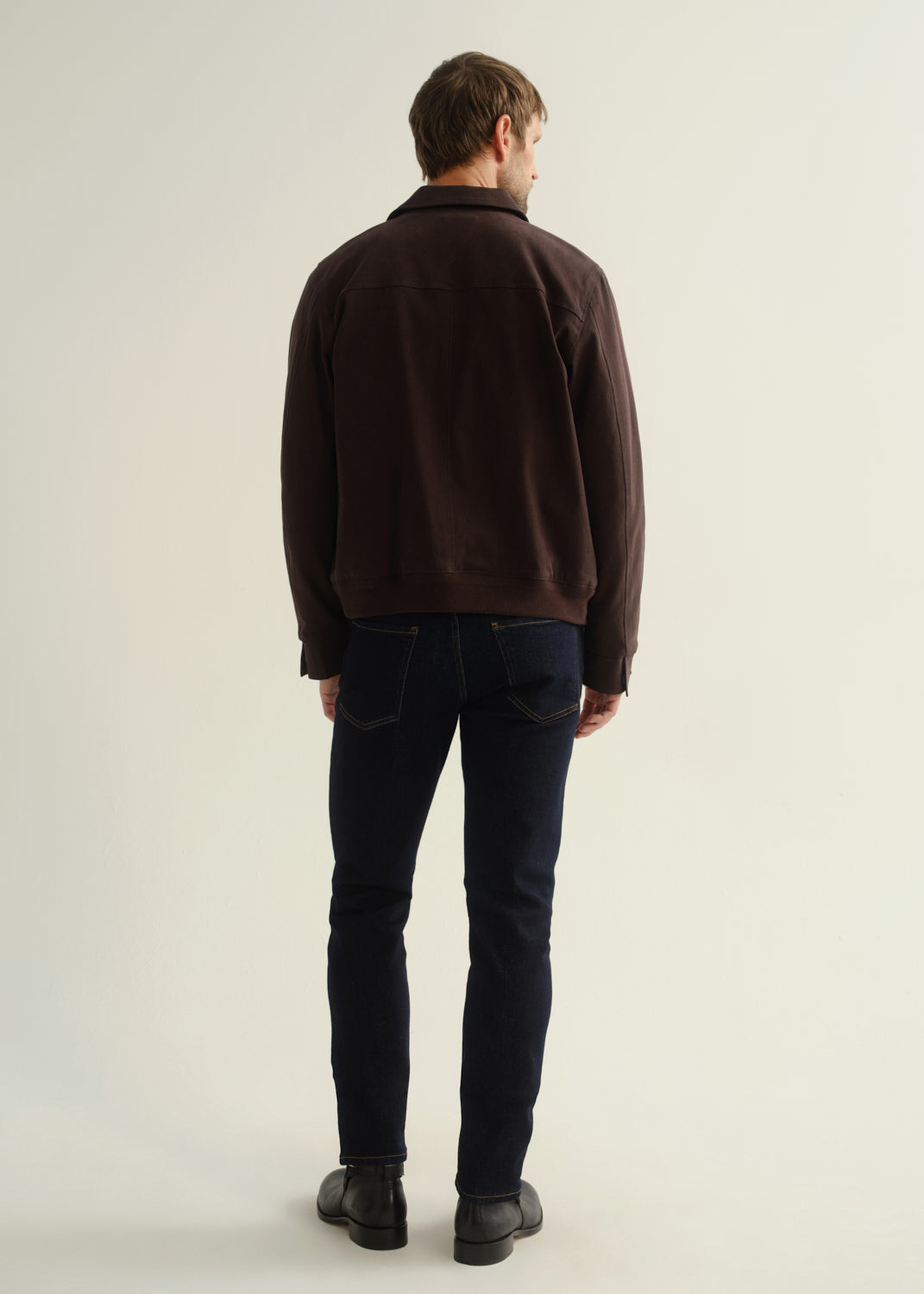 Person wearing a dark brown jacket, dark indigo jeans, and black leather boots, back full body view on a light grey background.