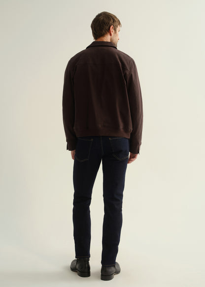 Person wearing a dark brown jacket, dark indigo jeans, and black leather boots, back full body view on a light grey background.