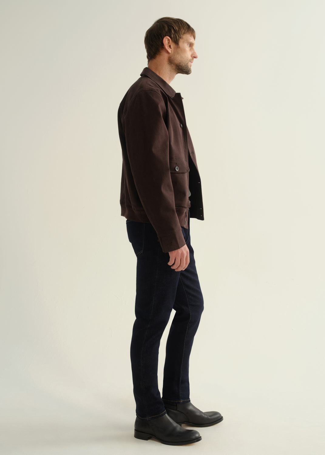 Person wearing a dark brown jacket, dark indigo jeans, and black leather boots, side full body view on a light grey background.