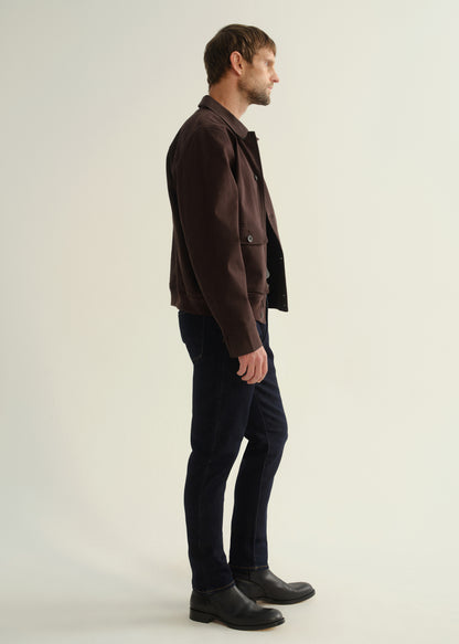 Person wearing a dark brown jacket, dark indigo jeans, and black leather boots, side full body view on a light grey background.