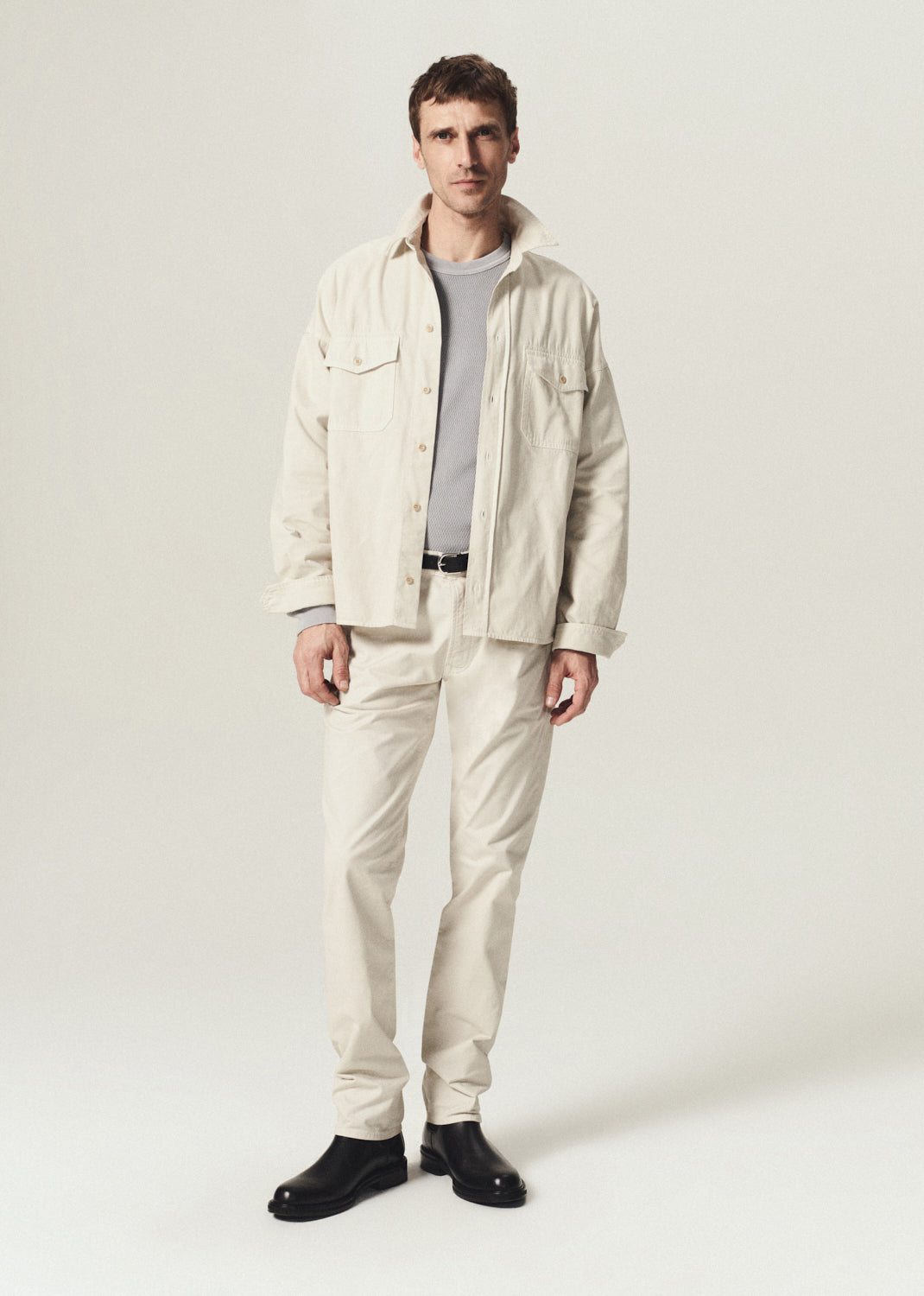 A man modeling a light beige linen jacket and matching trousers over a gray t-shirt and black shoes, standing against a plain light gray studio background.