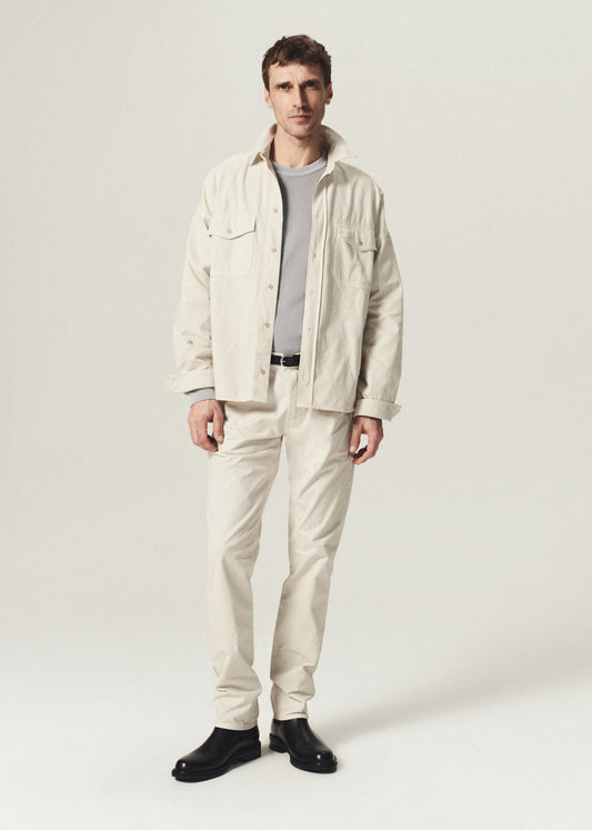 A man modeling a light beige linen jacket and matching trousers over a gray t-shirt and black shoes, standing against a plain light gray studio background.