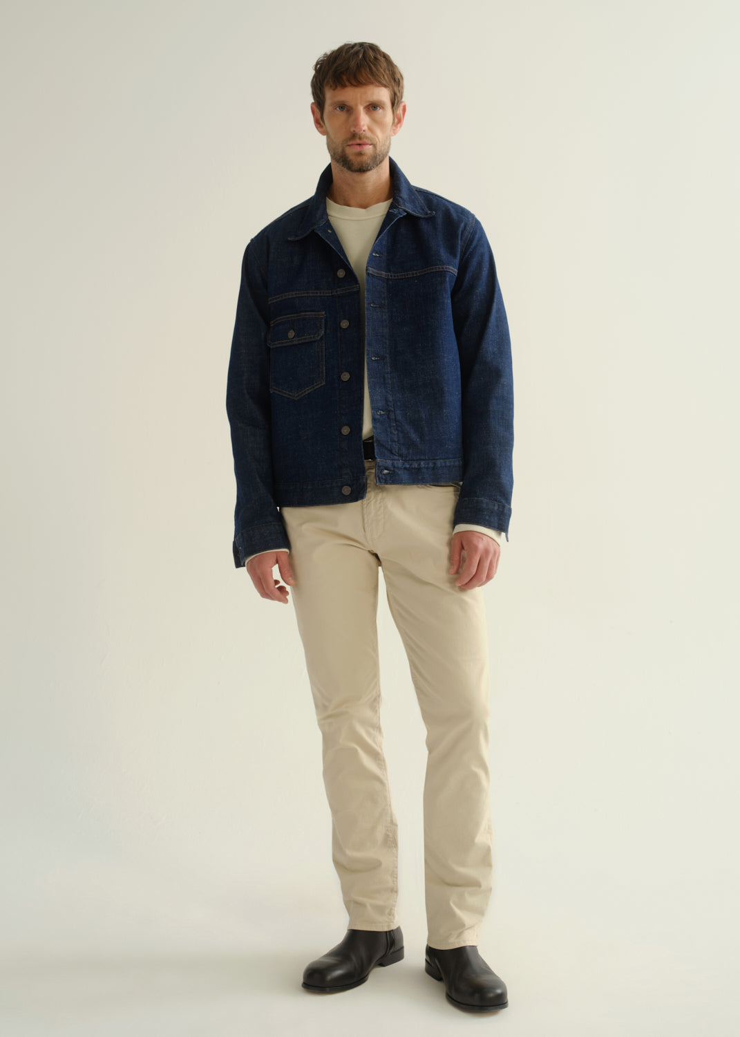 Person wearing a dark blue denim jacket, light beige pants, and black boots, front full body view on a light grey background.