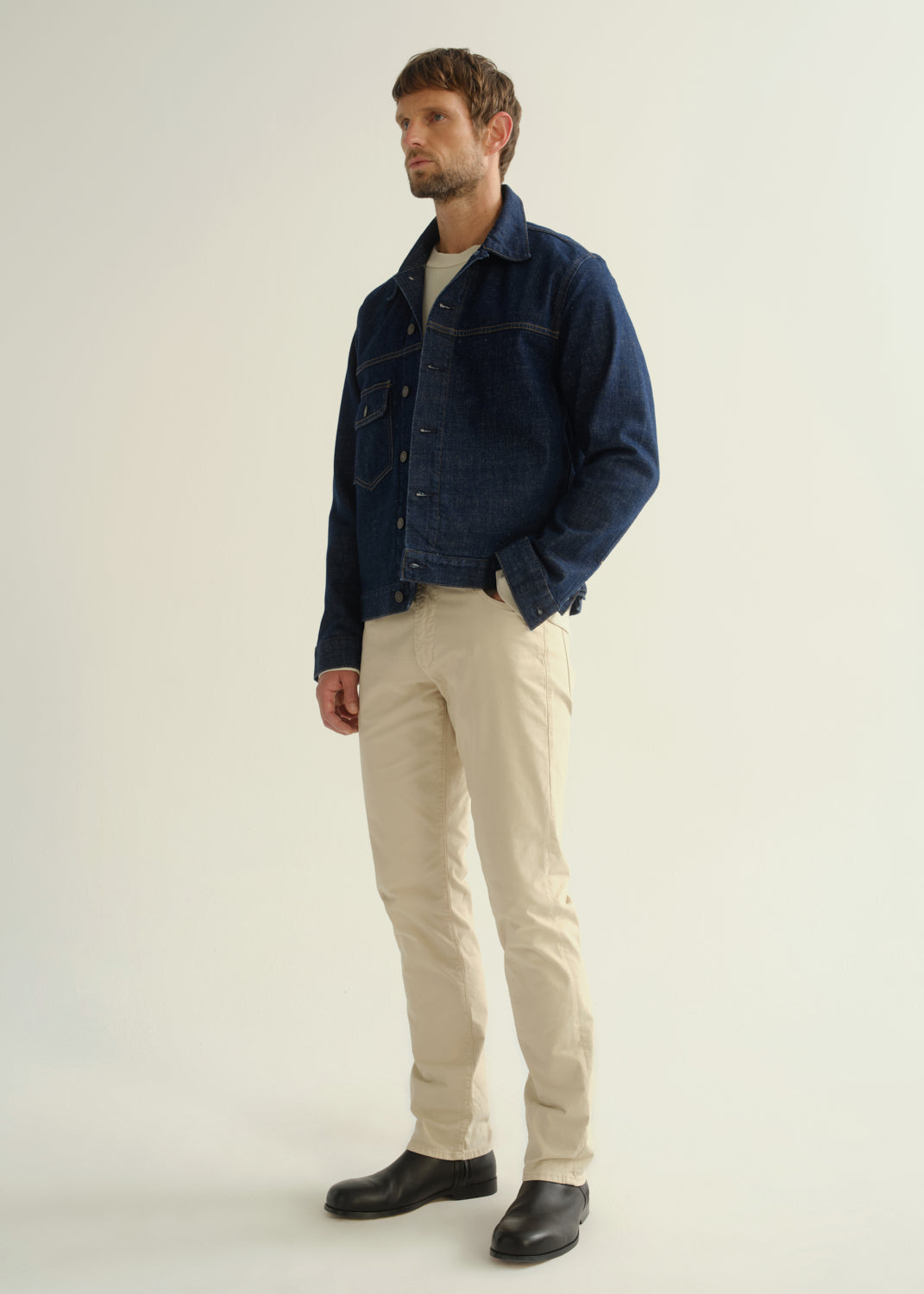 Person wearing a dark blue denim jacket, light beige pants, and black boots, angled front full body view on a light grey background.