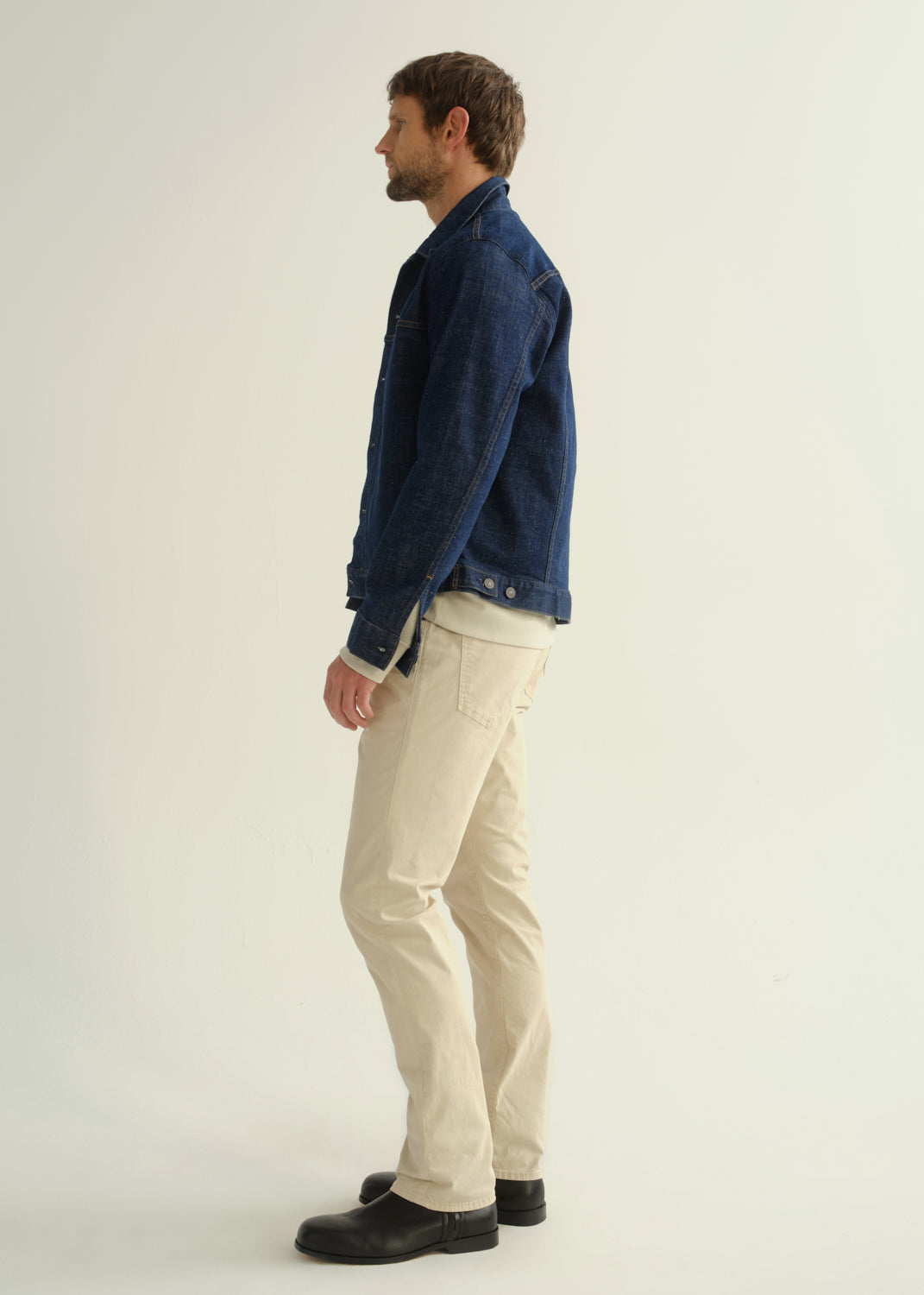 Person wearing a dark blue denim jacket, light beige pants, and black boots, side full body view on a light grey background.