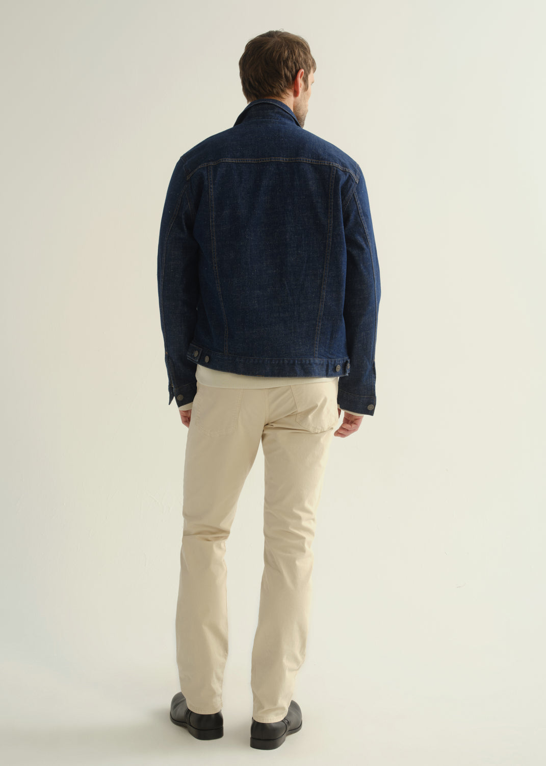 Person wearing a dark blue denim jacket, light beige pants, and black boots, back full body view on a light grey background.