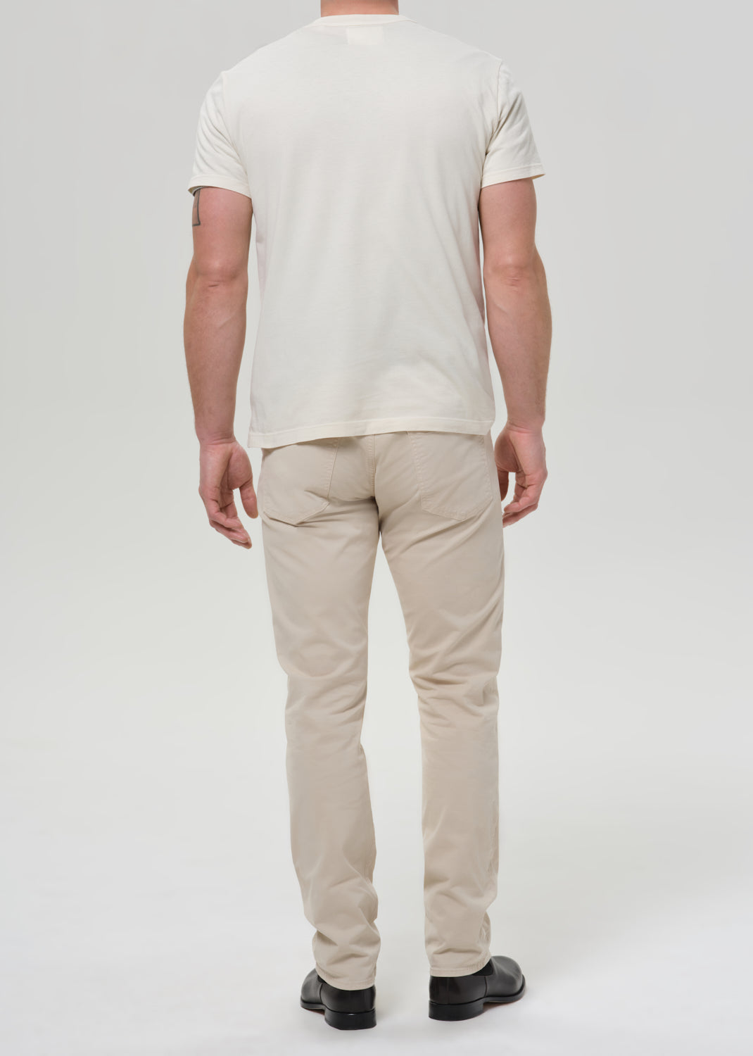 Person wearing cream straight leg pants with a white crewneck short sleeve tee viewed from the back on a grey background.