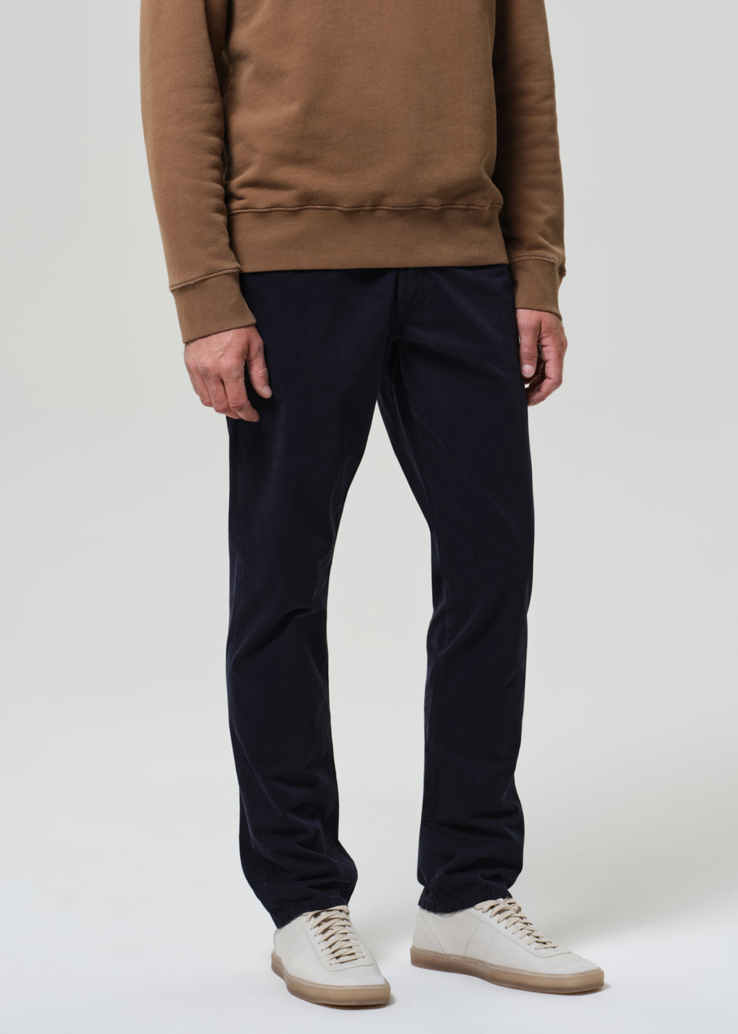 Person wearing a brown sweatshirt and black pants with white shoes on a plain background