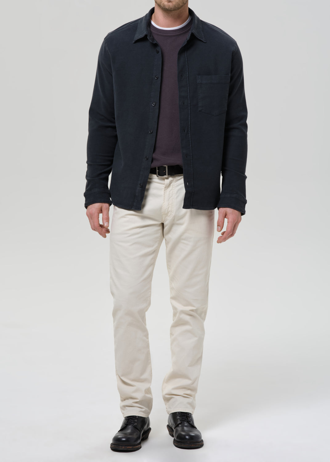 A man wearing a dark navy overshirt over a dark purple sweater, paired with off-white trousers and black leather shoes, standing against a grey background.