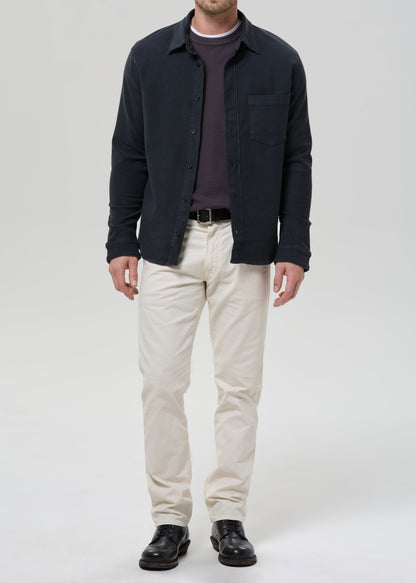 A man wearing a dark navy overshirt over a dark purple sweater, paired with off-white trousers and black leather shoes, standing against a grey background.