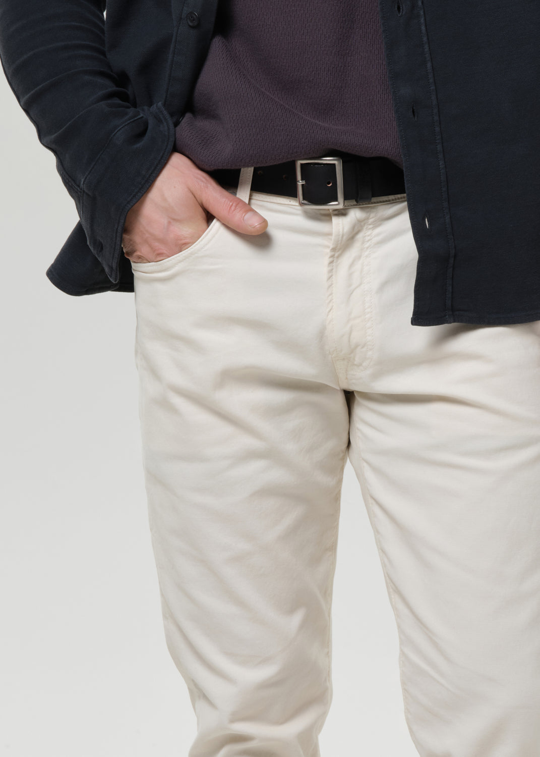 A close-up of a person wearing off-white or cream-colored chino pants with a black belt, styled with a dark purple shirt and a dark navy overshirt against a grey background.