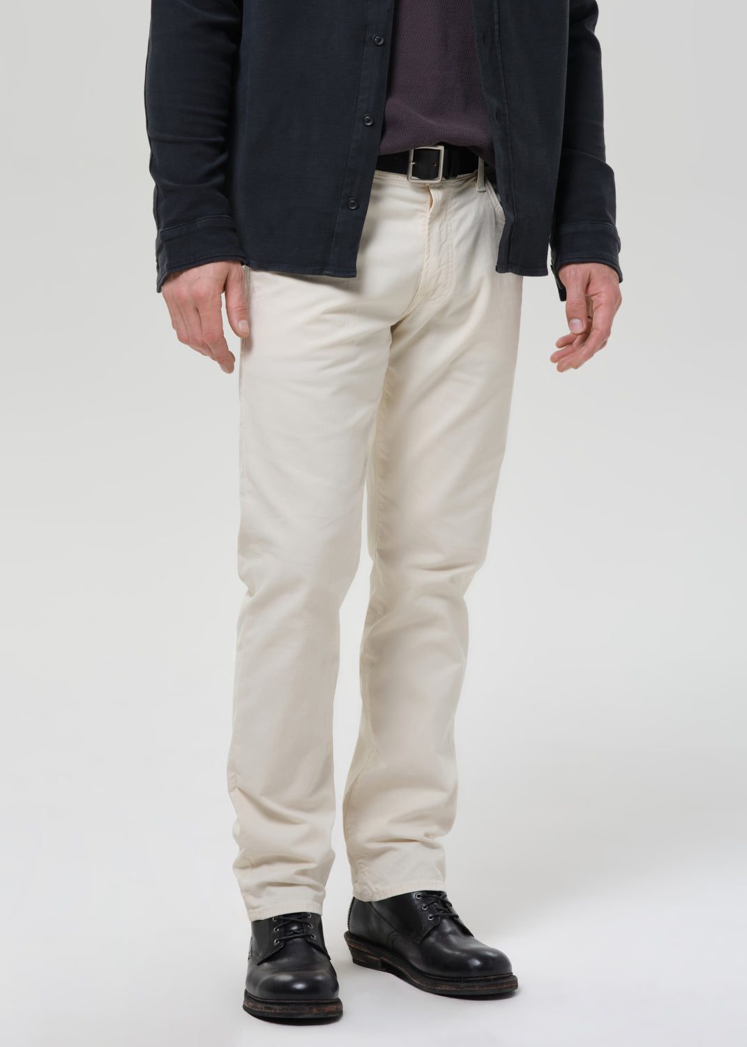 A person wearing off-white chino pants with a black belt, a dark navy overshirt, and black leather shoes, shown from the waist down on a grey background.