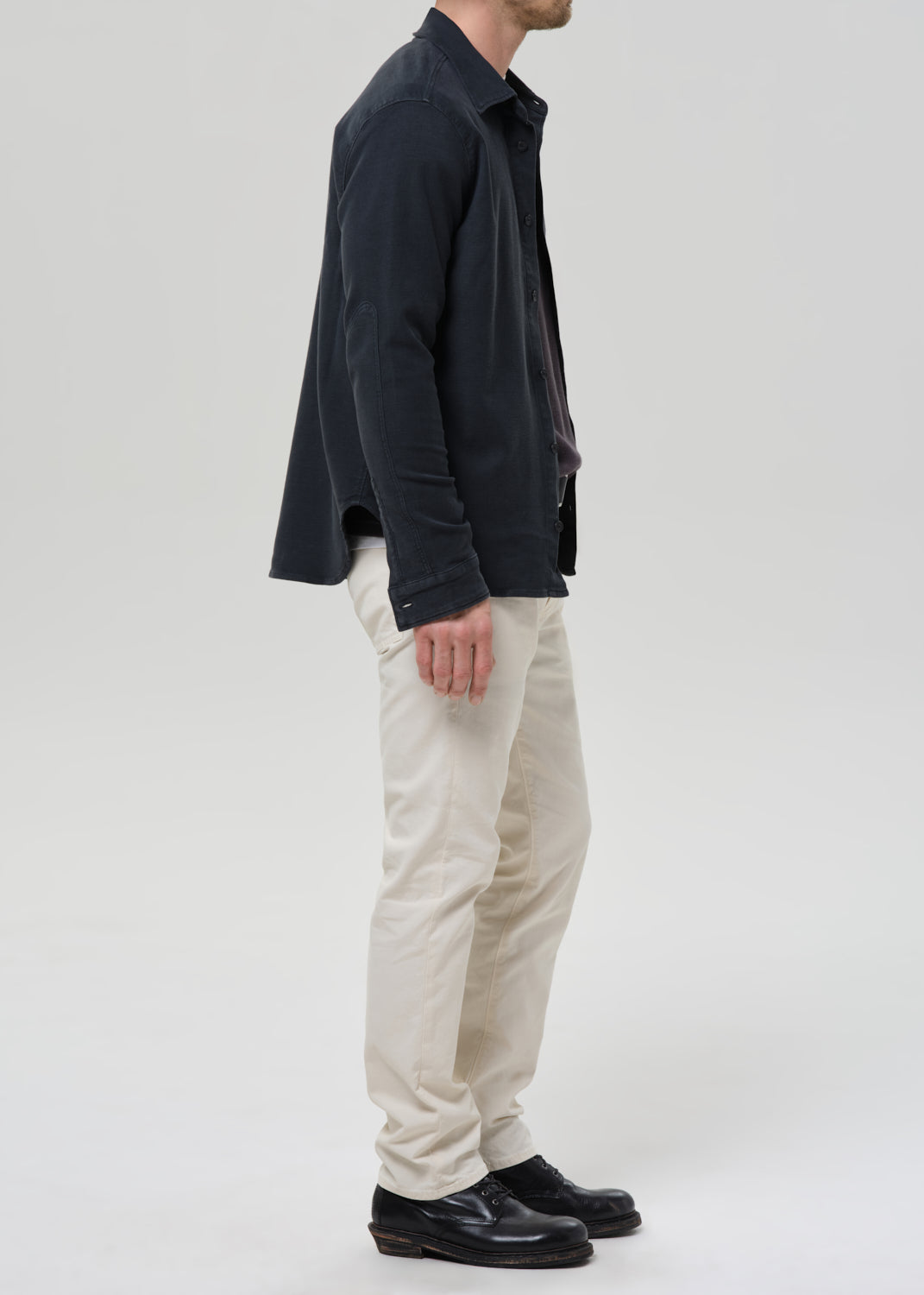 A man shown in profile wearing a dark navy overshirt, cream-colored trousers, and black leather shoes, photographed against a grey background.