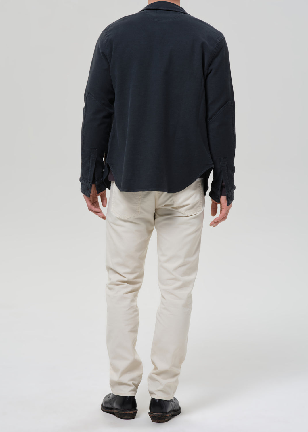 A man shown from the back wearing a dark navy long-sleeve button-up shirt, off-white chino pants, and black shoes, photographed against a grey background.