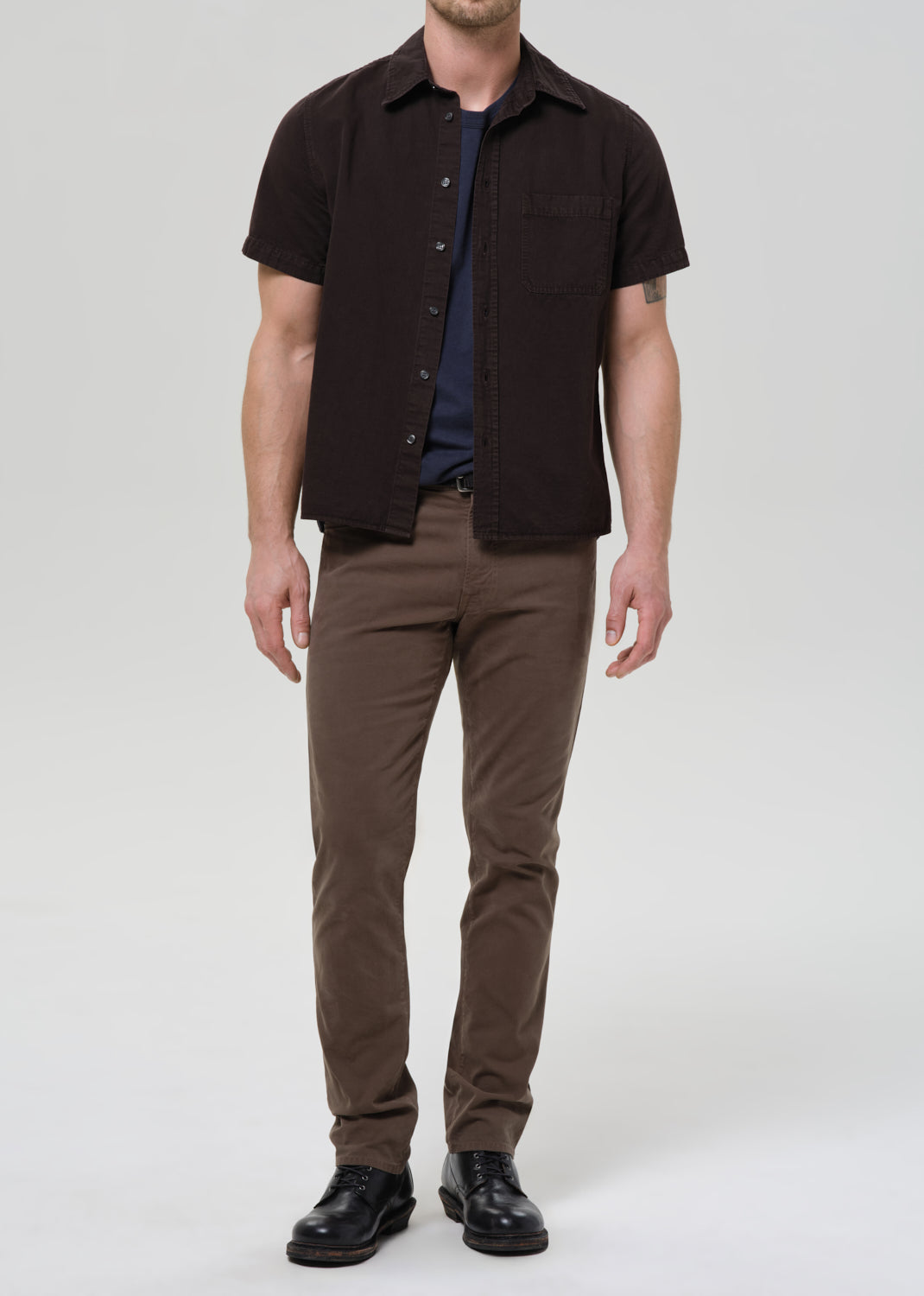 Man wearing a black short-sleeve shirt over a blue shirt, brown pants, and black shoes on a grey background