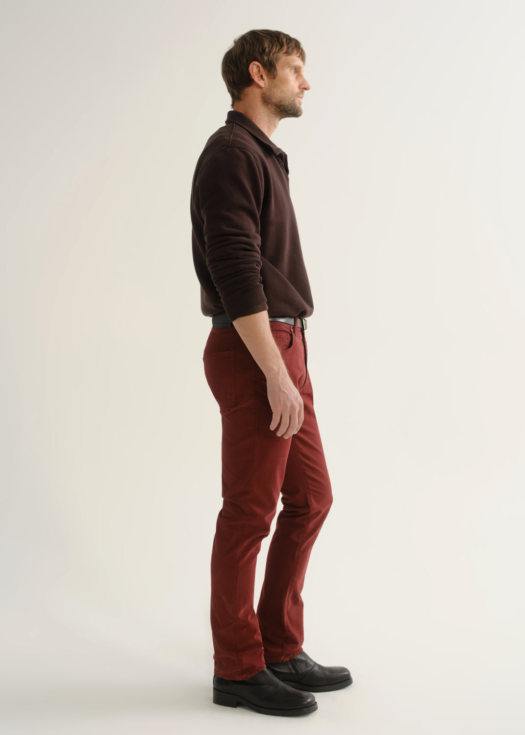 Person wearing a dark brown long sleeve top, burgundy pants, and black boots, side full body view on a light grey background.