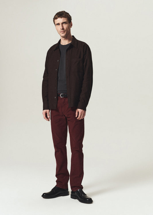 A man modeling a dark brown button-down shirt over a black t-shirt, paired with burgundy trousers and black leather shoes, standing against a light gray studio background.