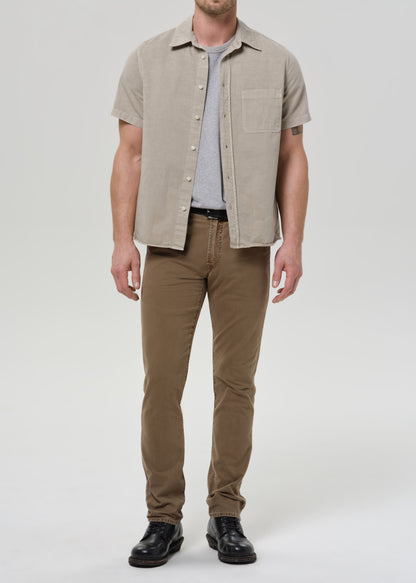 A man modeling a light beige short-sleeve button-up shirt over a grey t-shirt, paired with brown pants and black shoes, against a grey background.