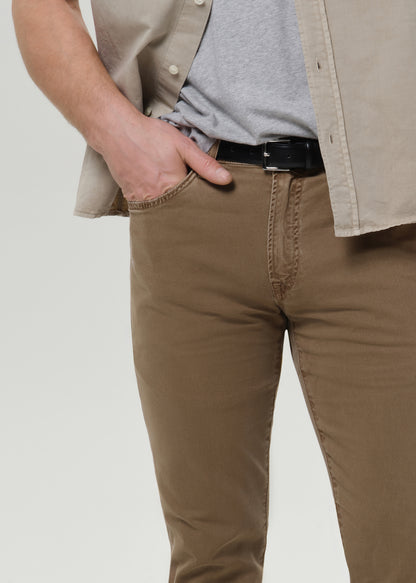 A close-up of a person wearing brown slim-fit jeans, styled with a black belt, a gray t-shirt, and a beige overshirt, set against a grey background.