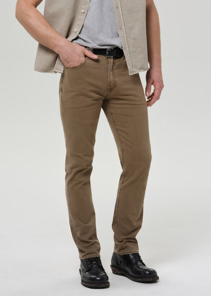 A person wearing slim-fit brown trousers, styled with a gray t-shirt, beige vest, black belt, and black leather shoes, photographed against a grey background.