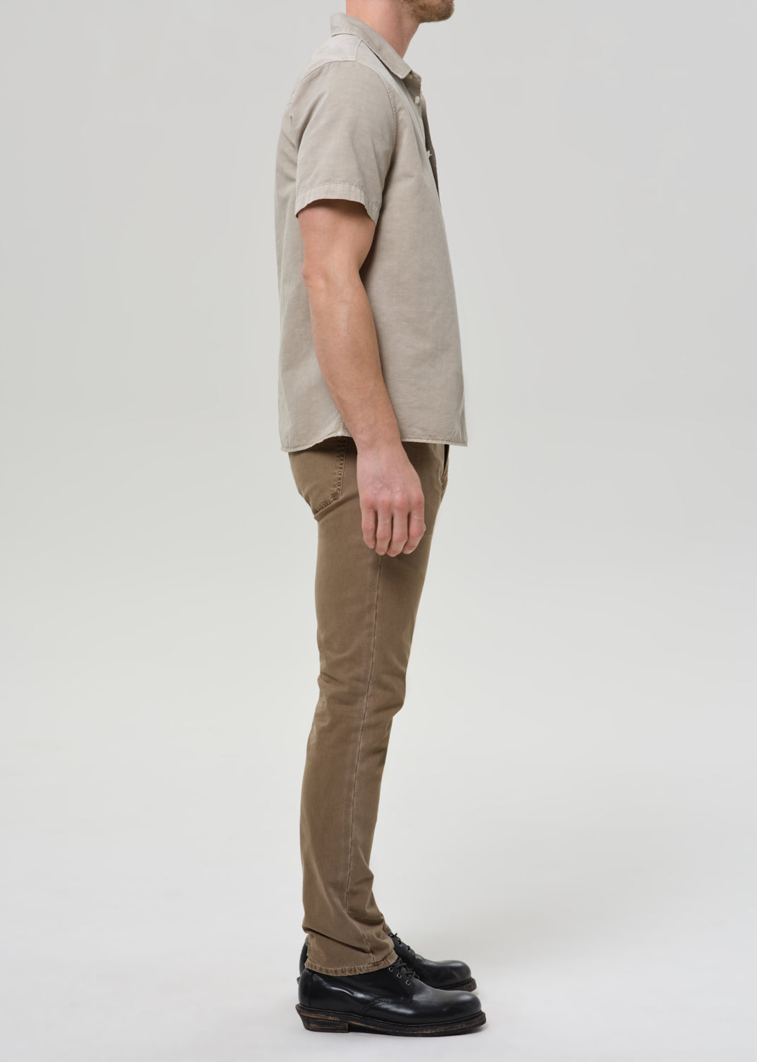 A man shown in profile wearing a light beige short-sleeve polo shirt, brown trousers, and black leather shoes against a grey background.