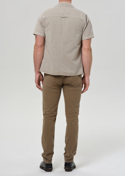 A man viewed from the back wearing a light beige short-sleeve button-up shirt and brown trousers, standing against a grey background.