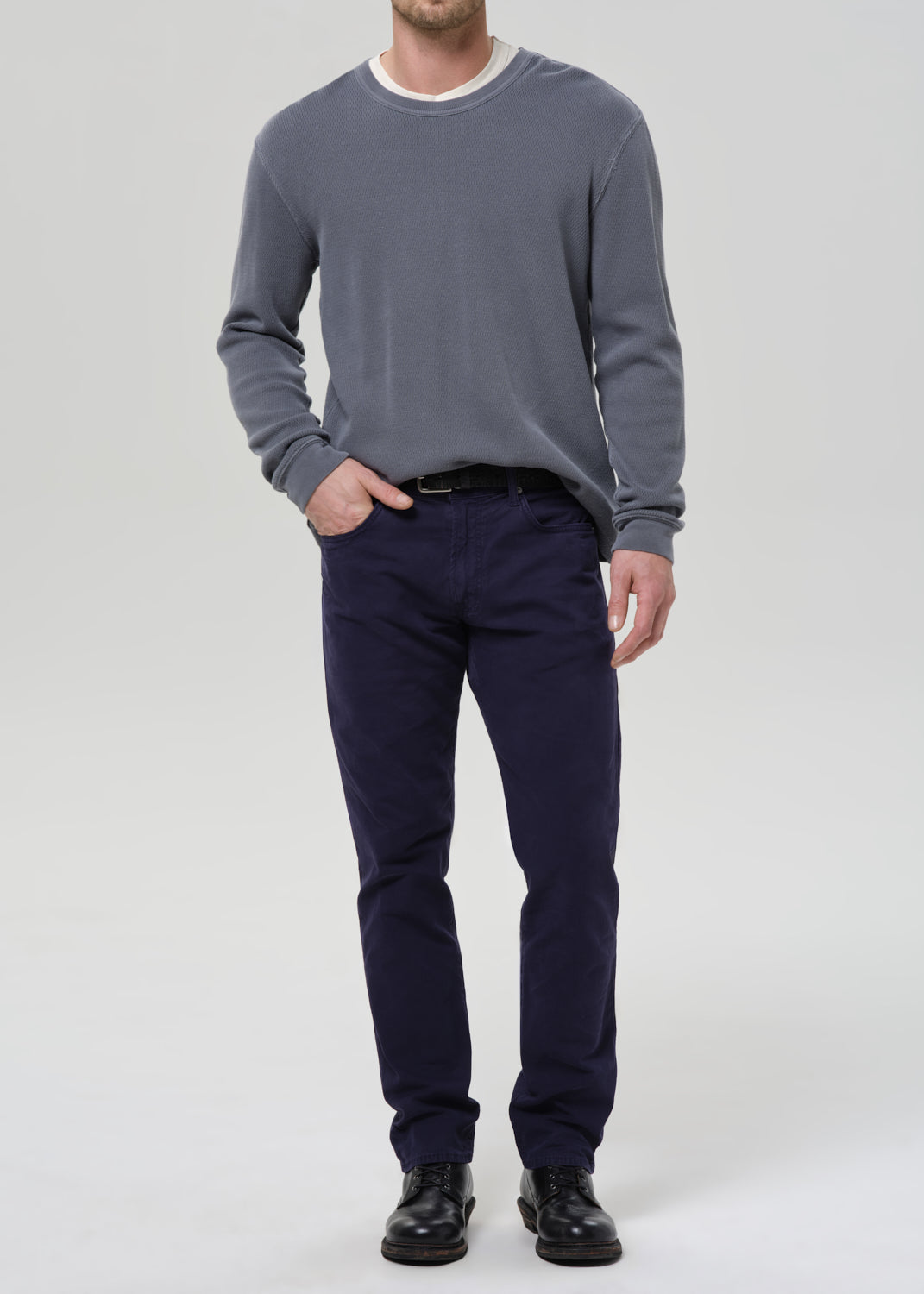 A man wearing a slate grey long-sleeve crewneck sweater, navy blue trousers, and black leather shoes, standing against a grey background.