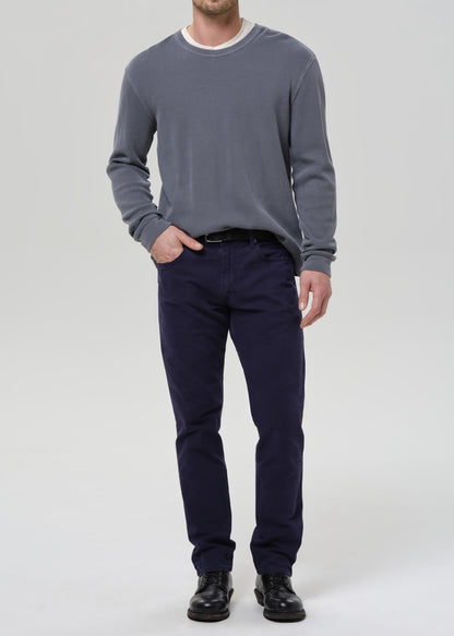A man wearing a slate grey long-sleeve crewneck sweater, navy blue trousers, and black leather shoes, standing against a grey background.