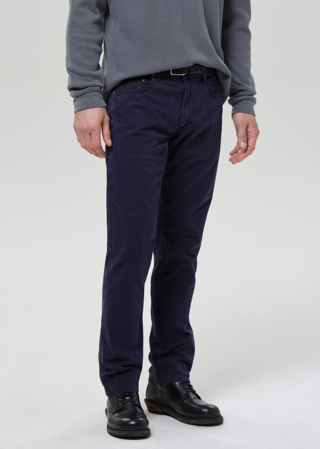 A person wearing dark navy slim-fit trousers, a grey long-sleeve top, and black leather shoes, standing against a grey background.