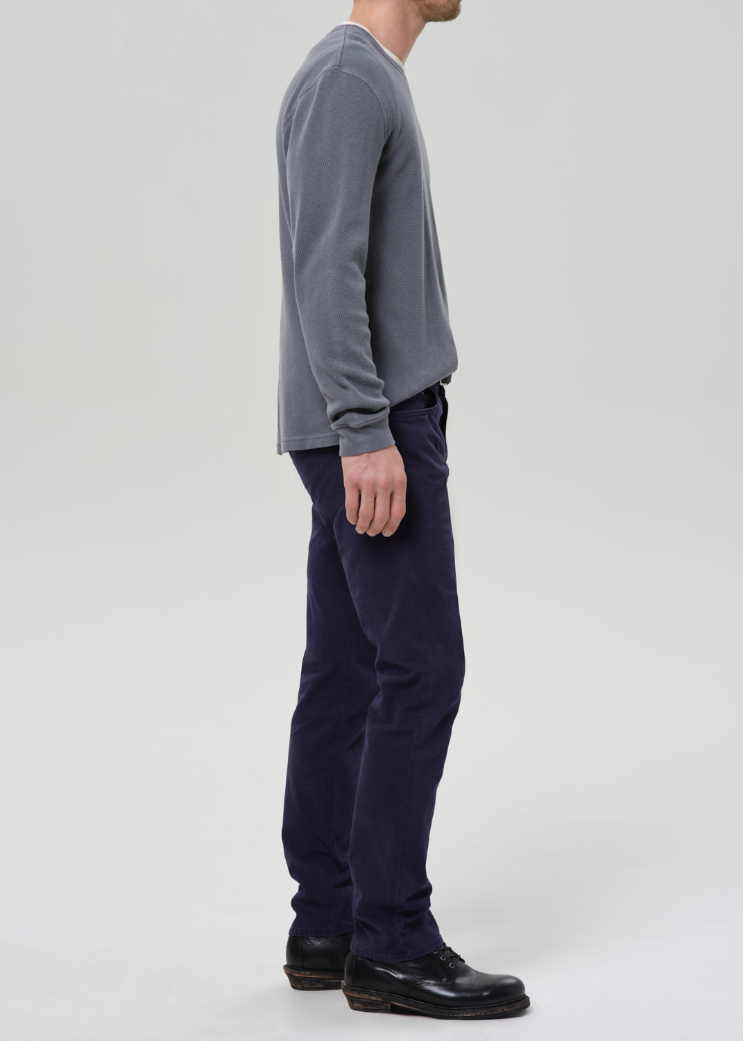 A man shown in profile wearing a grey long-sleeve shirt, dark navy pants, and black shoes, standing against a grey background.