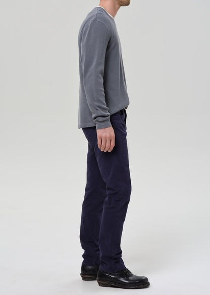 A man shown in profile wearing a grey long-sleeve shirt, dark navy pants, and black shoes, standing against a grey background.