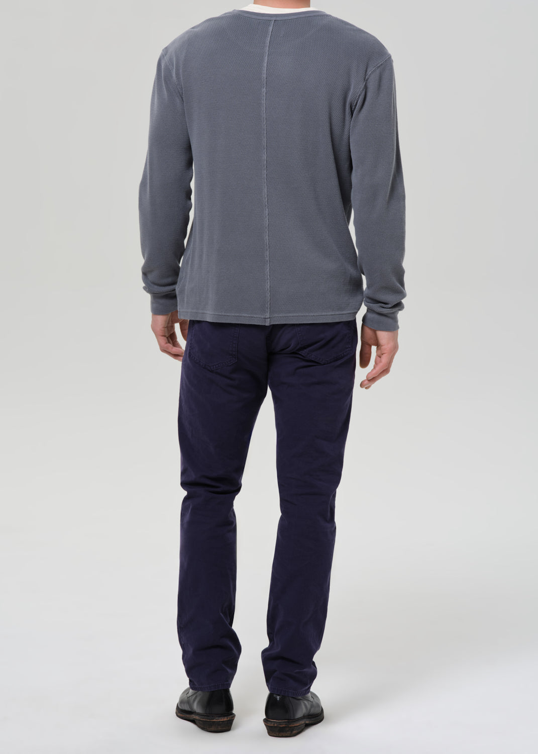 A man viewed from the back wearing a grey long-sleeve shirt and dark navy pants, standing against a grey background.