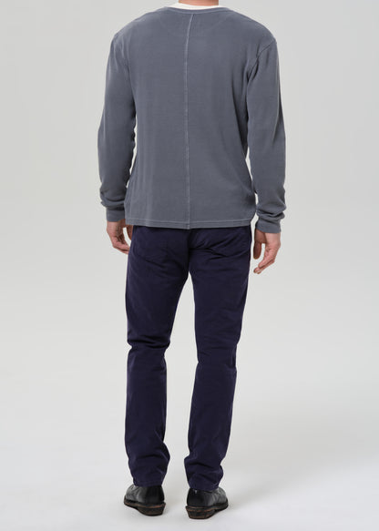 A man viewed from the back wearing a grey long-sleeve shirt and dark navy pants, standing against a grey background.