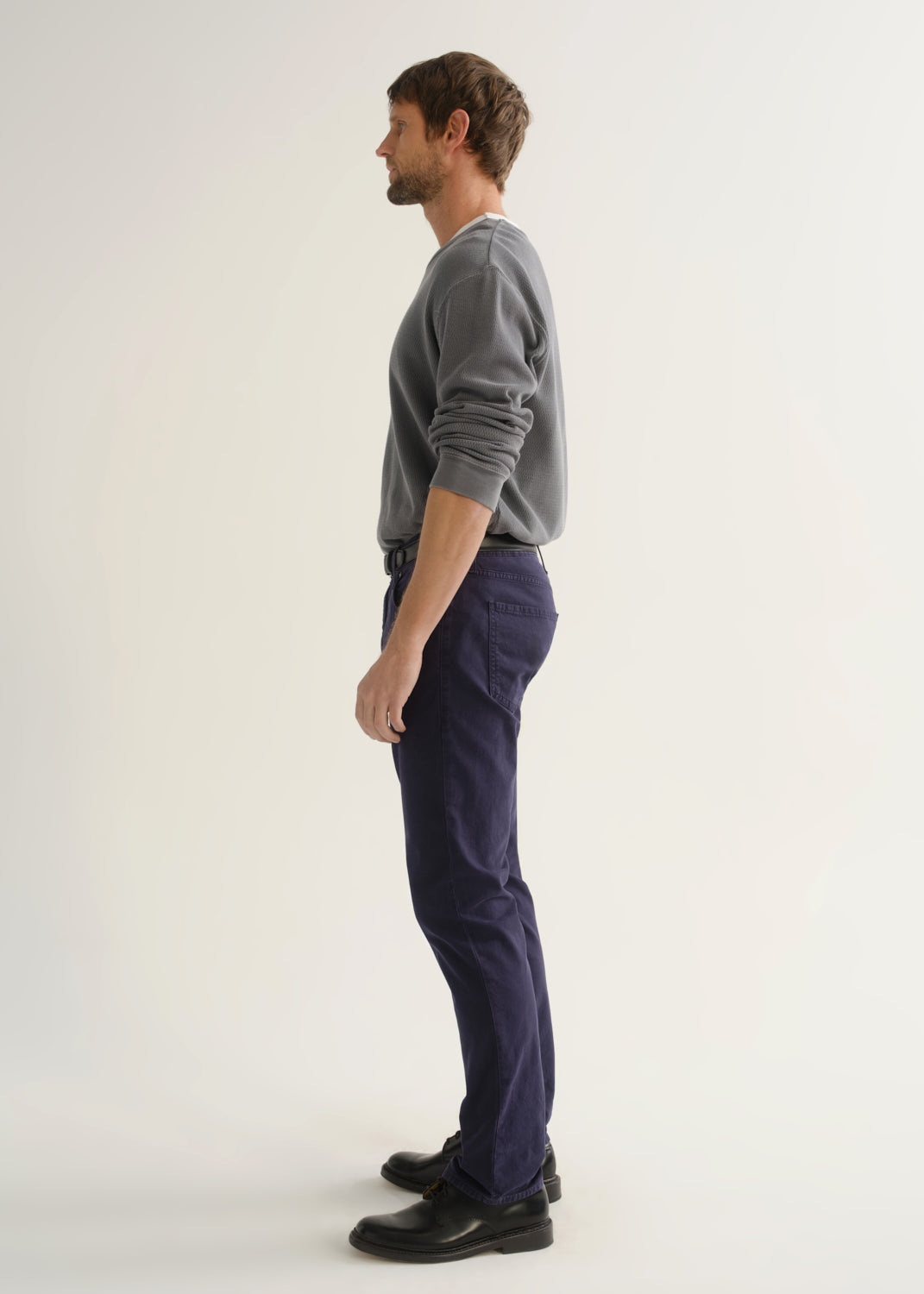 Person wearing a grey crewneck sweatshirt, dark navy pants, and black shoes, side full body view on a light grey background.
