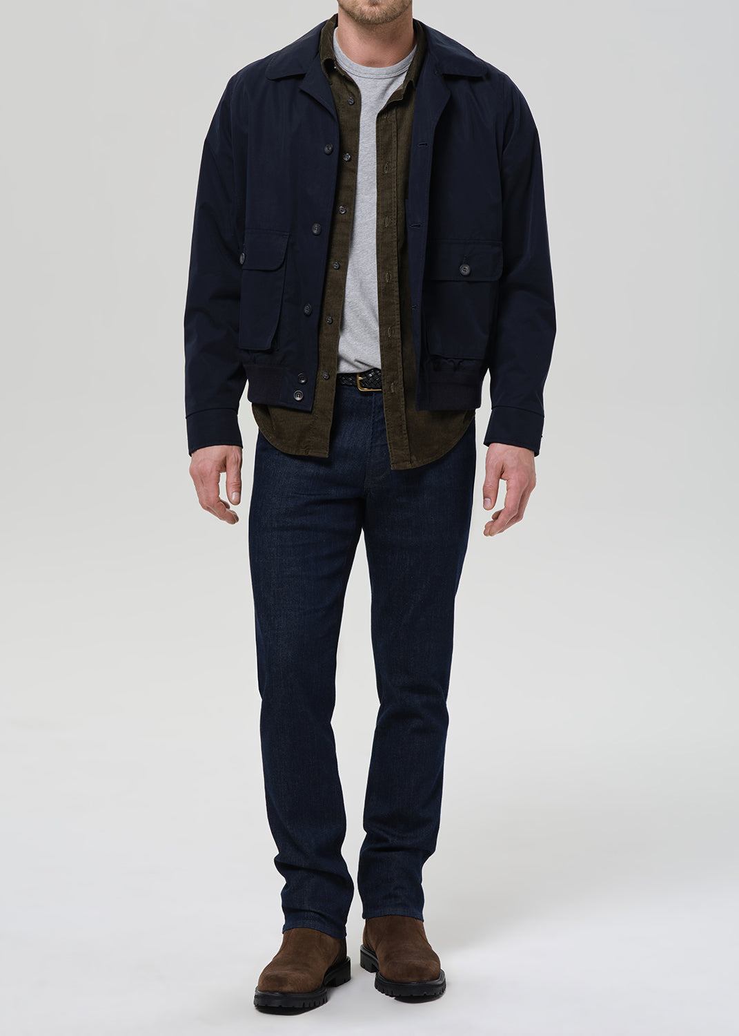 A man wearing a dark navy blue jacket over a brown corduroy shirt, a light gray t-shirt, dark blue jeans, and brown boots, standing against a light gray studio background.