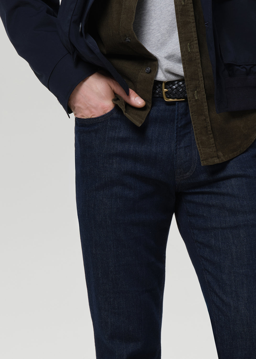 Close-up of a person wearing dark blue denim jeans, a gray t-shirt, an olive green overshirt, and a navy blazer, with one hand in the pocket of the jeans, against a light gray background.